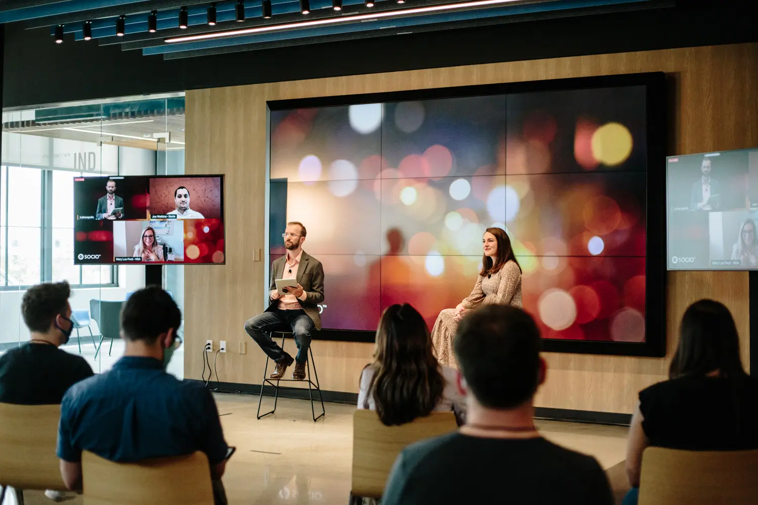 two people sitting in front of a video wall with two screens showing remote presenters